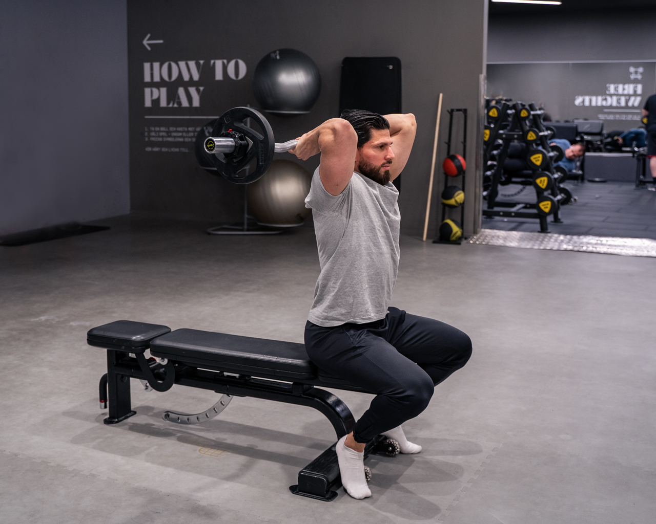 Person sitter på bänk och utför triceps extension bakom huvudet med skivstång – startläge med böjda armar