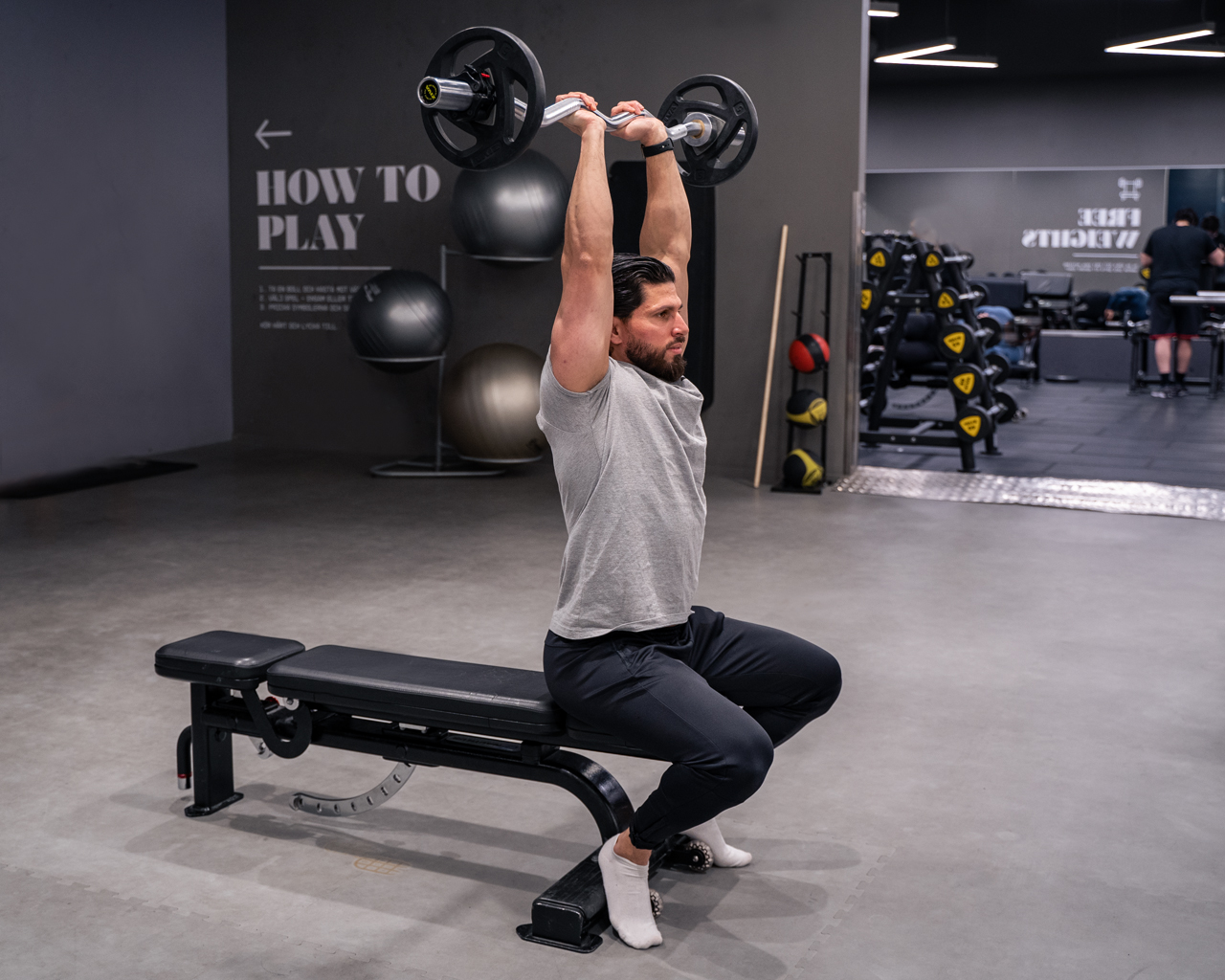 Man sitter på bänk och sträcker ut armarna rakt upp i triceps extension med skivstång – avslutande läge i övningen