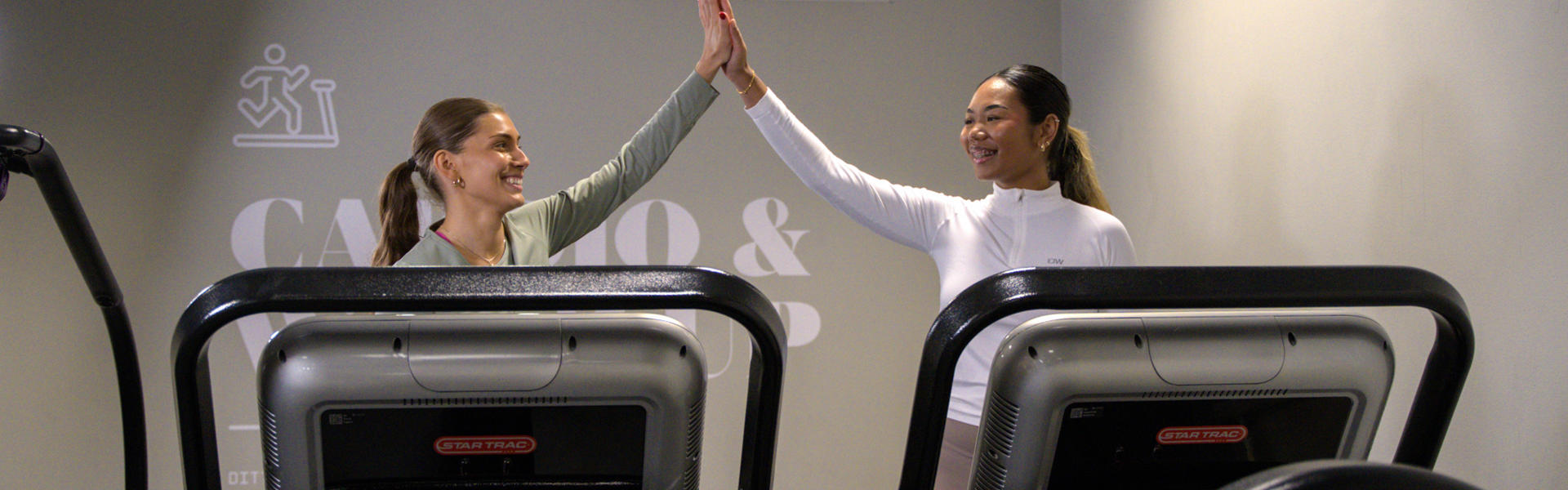 Två kvinnor gör high five med varandra när de står på sina löpband 